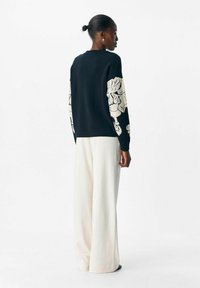 Suéter negro con patrones de punto blanco en las mangas, combinado con pantalones crema de corte holgado. El modelo está de espaldas, con el pelo recogido en un moño.