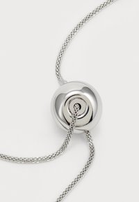 Un blocaj cilindric argintiu, realizat din metal lucios, cu un design în spirală, atașat la un fir subțire, împletit din argint.