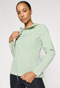 Chaqueta ligera de color verde claro con cierre de cremallera, capucha y mangas largas. Presenta un tejido texturizado y detalles reflectantes.