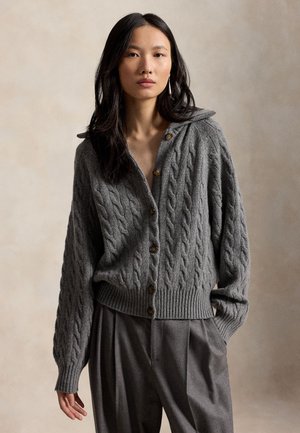 Femme portant un cardigan gris en maille torsadée boutonné et un pantalon gris ample, debout devant un fond neutre.
