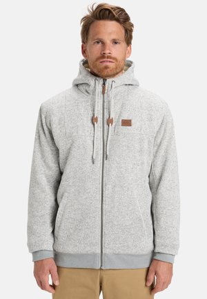 Man wearing light grey zip-up hoodie with drawstrings and beige pants, standing against a plain white background.