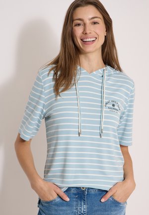 Femme souriante aux longs cheveux bruns portant un sweat à capuche à manches courtes rayé bleu clair et blanc et un jean bleu, les mains dans les poches.