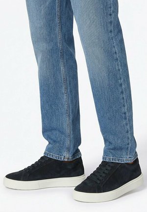 Osoba oblečená v modrých rifliach a čiernych semišových teniskách s bielymi podrážkami stojaca pred jednoduchým pozadím.