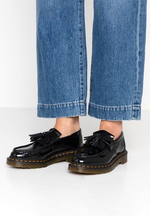 Glänzende schwarze Tasselloafers mit dicken braunen Sohlen, getragen zu hochgekrempelten blauen Denim-Jeans, gezeigt vom unteren Wadenbereich bis zu den Füßen auf einem weißen Boden.