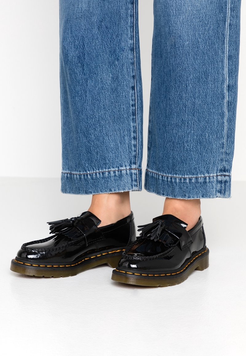 Mocassins noirs brillants à glands avec semelles épaisses marron, portés avec un jean en denim bleu retroussé, visibles de la partie inférieure du mollet jusqu’aux pieds sur un sol blanc.