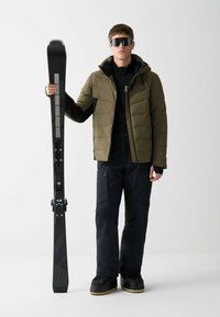 Snehové lyže s čiernym karbonovým designom. Model má na sebe olivovo zelenú prešívanú bundu, čierne termo tričko a námornícke nohavice. Look dopĺňajú slnečné okuliare.