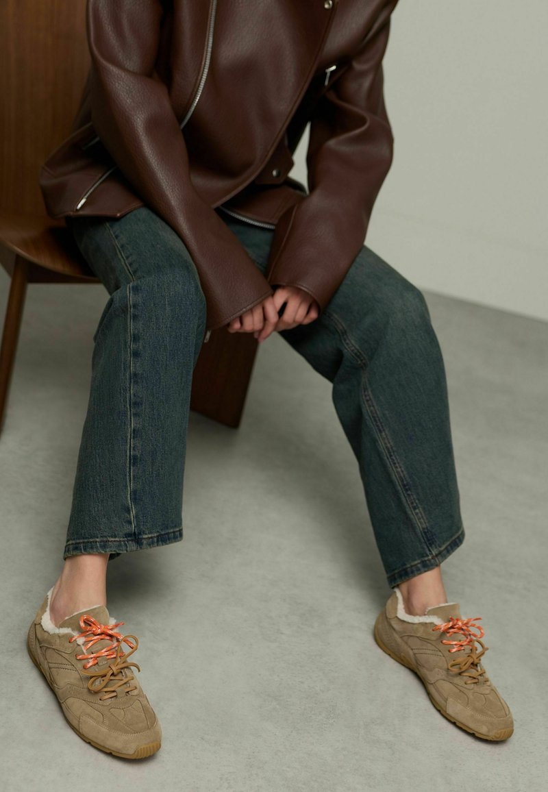 Beige suede sneakers with orange laces, featuring a fluffy trim, paired with dark jeans and a brown leather jacket, seated on a wooden chair.