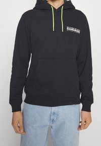 Sudadera negra con cordones de neón verde, bolsillo frontal, logo impreso en el pecho izquierdo y puños y dobladillo acanalados, llevada con jeans azul claro.