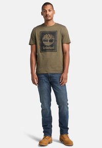 T-shirt in cotone verde oliva con un design del logo Timberland ad albero nero all'interno di un quadrato. Indossato con jeans denim blu e stivali beige.