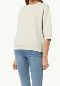 Vrouw draagt een roomkleurige oversized sweatshirt met driekwart mouwen en lichtblauwe strakke jeans, staand tegen een witte achtergrond.