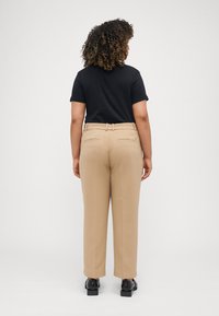 Beige tilpassede bukser med rette ben, med belte i midjen, to lommer og en glatt stofftekstur. Svarte sko er iført.