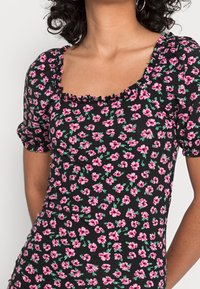 Czarna bluzka w kwiaty z krótkimi bufiastymi rękawami, kwadratowym dekoltem i falbaną na krawędzi. Wzór z małymi różowymi kwiatami i zielonymi liśćmi.