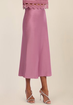 Frau trägt einen midilangen pinkfarbenen Satinsrock mit Spitzenbesatz am Saum, ein Top und silberne High-Heels mit Riemchen, steht vor einem einfarbigen Hintergrund.