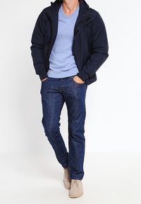 Veste zippée marine sur un pull col V bleu clair, jean denim foncé et chaussures beiges. Tenue décontractée superposée avec des couleurs contrastantes.