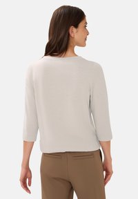 Light gray, three-quarter sleeve top with a round neckline, made from a textured fabric, paired with brown trousers featuring back pockets.