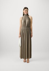 Pleated halter neck dress in a muted taupe color, with a smocked waist and flowing skirt. Paired with a white handbag and strappy heels.