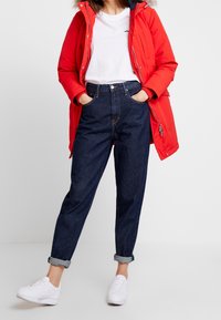 Rode donsjack met een capuchon met bontvoering, wit shirt met korte mouwen, donkerblauwe jeans met opgerolde pijpen en witte sneakers.