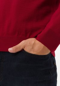 Pull en maille rouge avec poignets et ourlet côtelés, porté avec un jean en denim foncé. Main reposant dans la poche, mettant en valeur la texture du tissu et le contraste des couleurs.