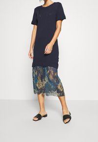 Granatowa sukienka z krótkim rękawem i przezroczystą, midi spódnicą w kwiatowy wzór, zestawiona z czarnymi klapkami. Swobodny, zwiewny krój z warstwowanymi teksturami.