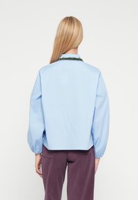 Lyseblå bluse med puffede �ærmer og cropped design. Har en grøn dekorativ kant rundt om kraven. Glat bomuldsstof.