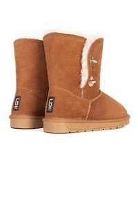 Botines de ante marrón con un suave forro de piel blanca, que cuentan con dos botones decorativos y una suela texturada de color marrón claro. Parche con el logo G en la parte trasera.