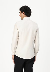 Lys beige langermet skjorte med strukturert krage, skreddersydd passform, knappkant og subtil tekstur. Brukt med svarte bukser.
