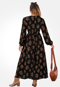 Černé květinové šaty se zlatými botanickými vzory. Dlouhé rukávy a stažený pas. Hnědá kabelka přes rameno a kotníkové boty dotvářejí vzhled.
