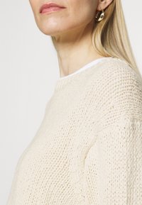 Crème kleurige gebreide trui met een textuurpatroon en ronde hals, gelaagd over een wit T-shirt; zichtbare gouden creolen.