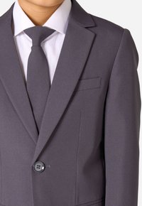 Graues Sakko mit Revers, Einzelknopf-Detail und einer aufgesetzten Tasche. Getragen über einem weißen Hemd und einer grauen Krawatte.