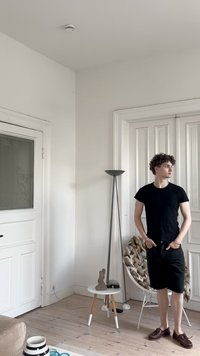 Černé tričko, černé šortky a hnědé boty nosí osoba v světlé místnosti s bílými stěnami, dřevěnou podlahou a moderními dekoračními prvky.