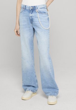 Personne portant un jean bleu clair ample avec une chaîne de perles sur la poche droite et des baskets blanches, sur un fond uni.