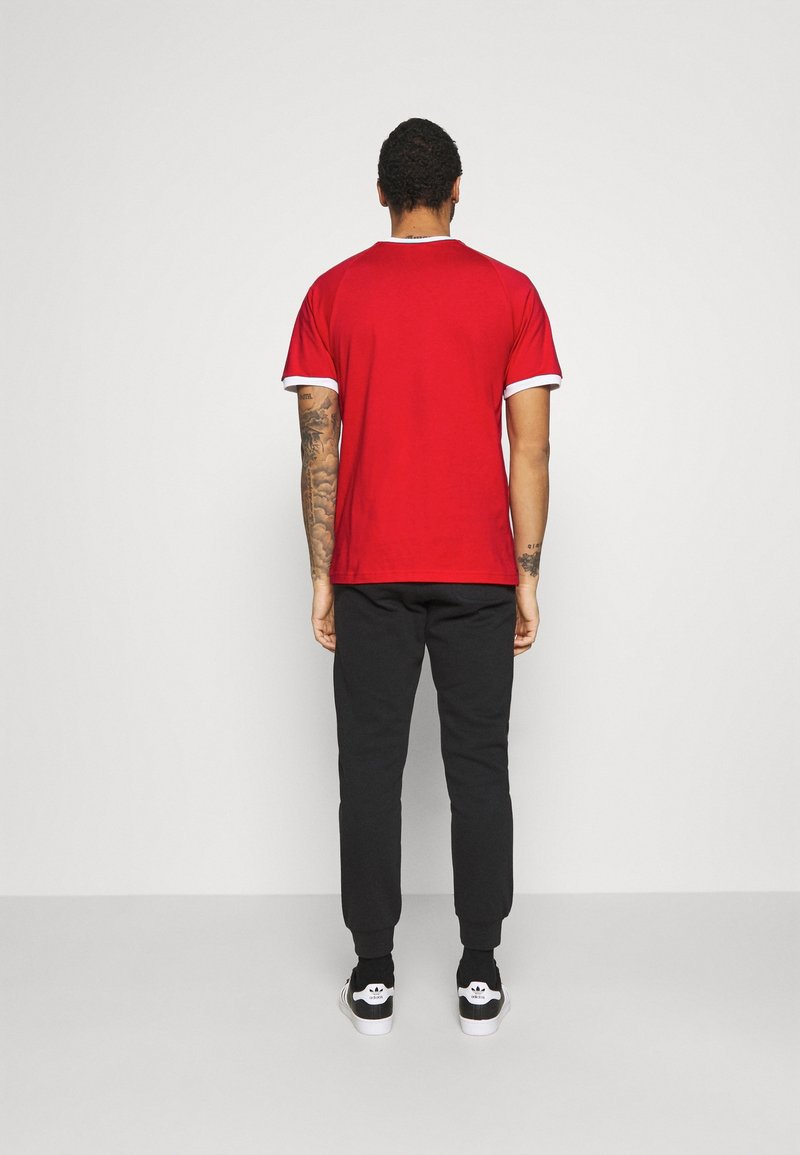 Camiseta roja con ribete blanco en el cuello, combinada con pantalones jogger negros. Tatuajes visibles en los brazos, usando zapatos negros con acentos blancos.
