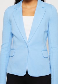 Jasnoniebieska marynarka wykonana z teksturowanej tkaniny, z pojedynczym zapięciem na guzik, klapami z wycięciami oraz dwiema przednimi kieszeniami.