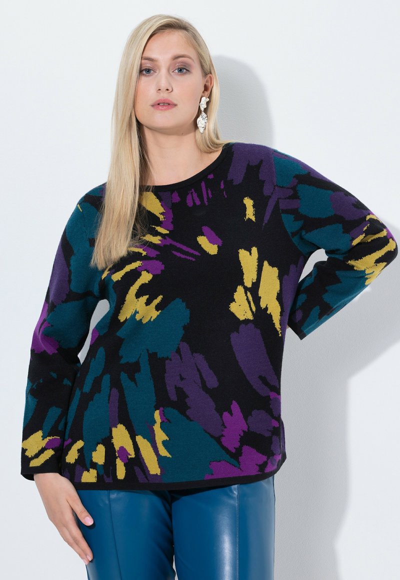 Donna bionda che indossa un maglione a fantasia viola, teal e giallo e pantaloni di pelle blu, in piedi con una mano sui fianchi contro un muro bianco.