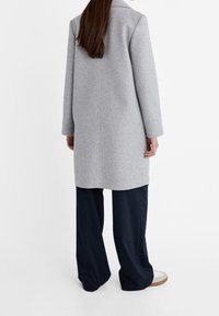 Grijze wollen jas, halflang met lange mouwen, strakke lijnen, detail in de achternaad, gedragen over een marineblauwe wijde broek en witte sneakers.