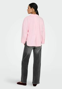 Femme avec queue de cheval noire portant une chemise ample rose pâle, un jean large noir délavé et des ballerines rouge foncé, debout de dos.