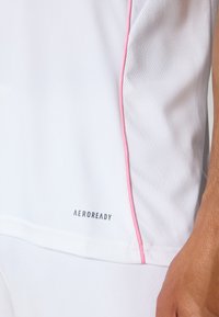 Fehér sportpóló, rózsaszín díszítéssel az oldalon. Légáteresztő anyagból készült, az alsó részén 'AEROREADY' logóval. Texturált hálós panel látható.