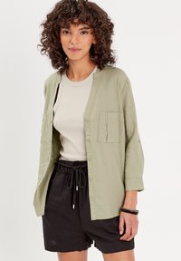 Chemise boutonnée vert clair avec deux poches avant, portée sur un débardeur beige, associée à un short noir avec taille à cordon.