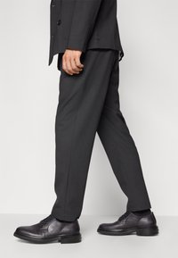 Mörkgrå läderskor med snörning och rund tå, minimal sömnad och en tjock svart sula, tillsammans med skräddarsydda mörka byxor.