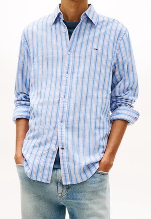 Homme portant une chemise à rayures bleu clair boutonnée avec les manches retroussées et un jean bleu clair, mains dans les poches, sur fond blanc.