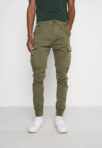 Olivengrønne cargo-bukser med flere lommer, tapered fit, elastiske manchetter og knaplukning, parret med hvide sneakers.