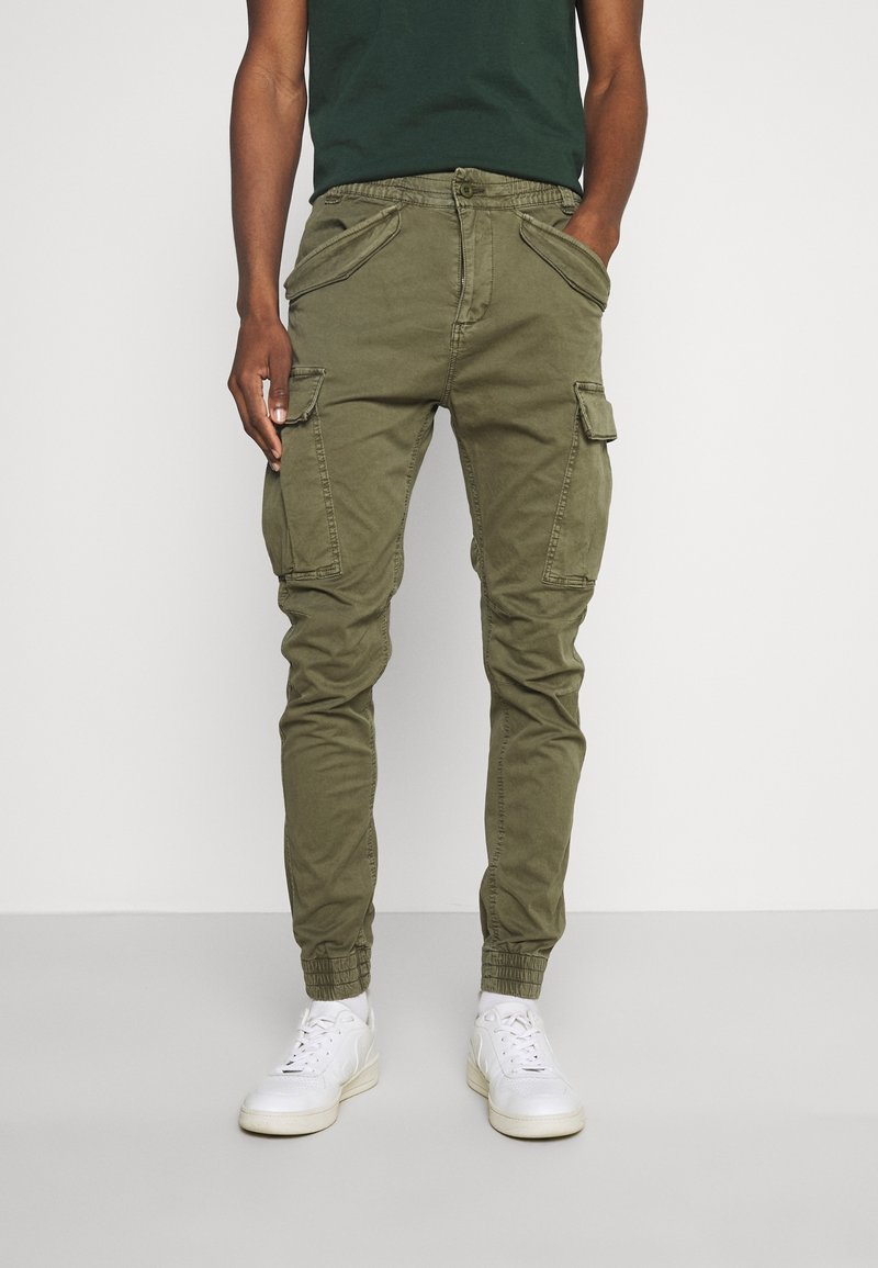 Olivengrønne cargo-bukser med flere lommer, tapered fit, elastiske manchetter og knaplukning, parret med hvide sneakers.