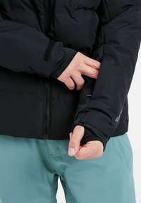 Schwarze isolierte Jacke mit durchgehendem Reißverschluss, verstellbaren Manschetten und glatter Textur, getragen mit hellblauen Hosen.