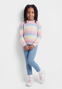 Μικρή κοπέλα με πλεγμένα μαλλιά, φορούν πουλόβερ με ριγέ παστέλ χρώματα, ανοιχτόχρωμο μπλε τζιν, άσπρες κάλτσες με ροζ καρδιές και λευκά αθλητικά παπούτσια με ροζ κορδόνια.