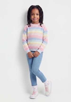 Jong meisje met gevlochten haar, een pastelgestreepte trui, lichtblauwe spijkerbroek, witte sokken met roze hartjes en witte sneakers met roze veters.