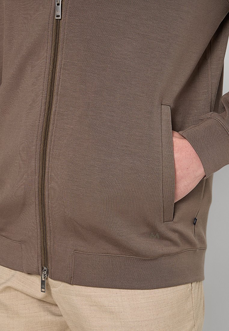Brązowa kurtka zapinana na zamek z ręką w bocznej kieszeni, w połączeniu z beżowymi spodniami, zaprezentowana na osobie na neutralnym tle.