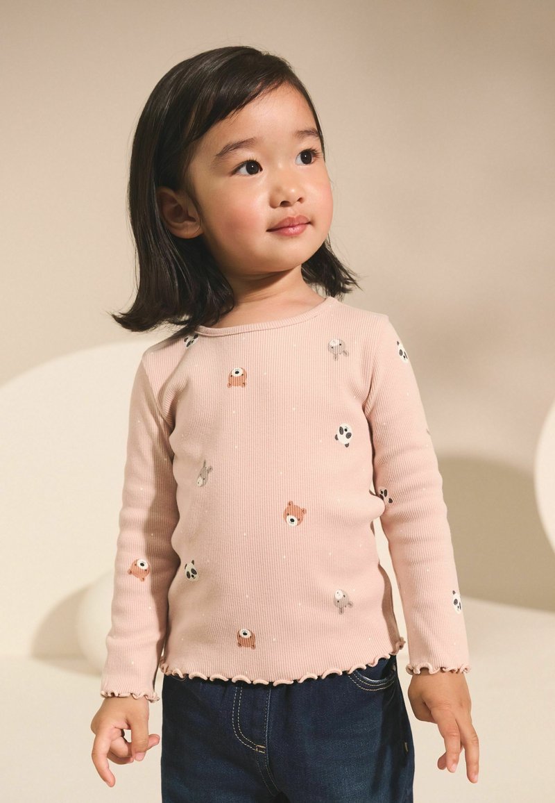 Chemise à manches longues rose clair avec texture côtelée, ornée de petits imprimés d'animaux et d'un ourlet festonné. Associée à un jean bleu foncé.