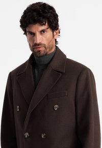 Brown wool double-breasted coat features large buttons, peaked lapels, and a chest pocket. Worn over a dark turtleneck sweater.