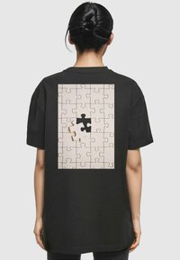 Sort T-shirt med et stort puslespilsdesign p�å ryggen, bestående af beige puslespilsbrikker og et enkelt sort brik, der passer ind i designet.