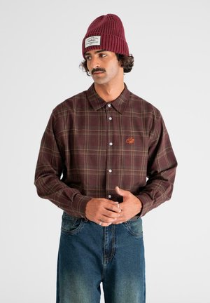 Man in maroon plaid button-up shirt and knitted maroon beanie with patch, wearing blue jeans, standing against white background.